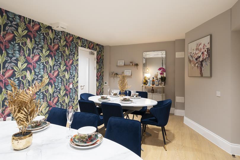 A modern dining room with a white oval table, blue velvet chairs, tropical leaf wallpaper, light wood floor, and a bar cart w