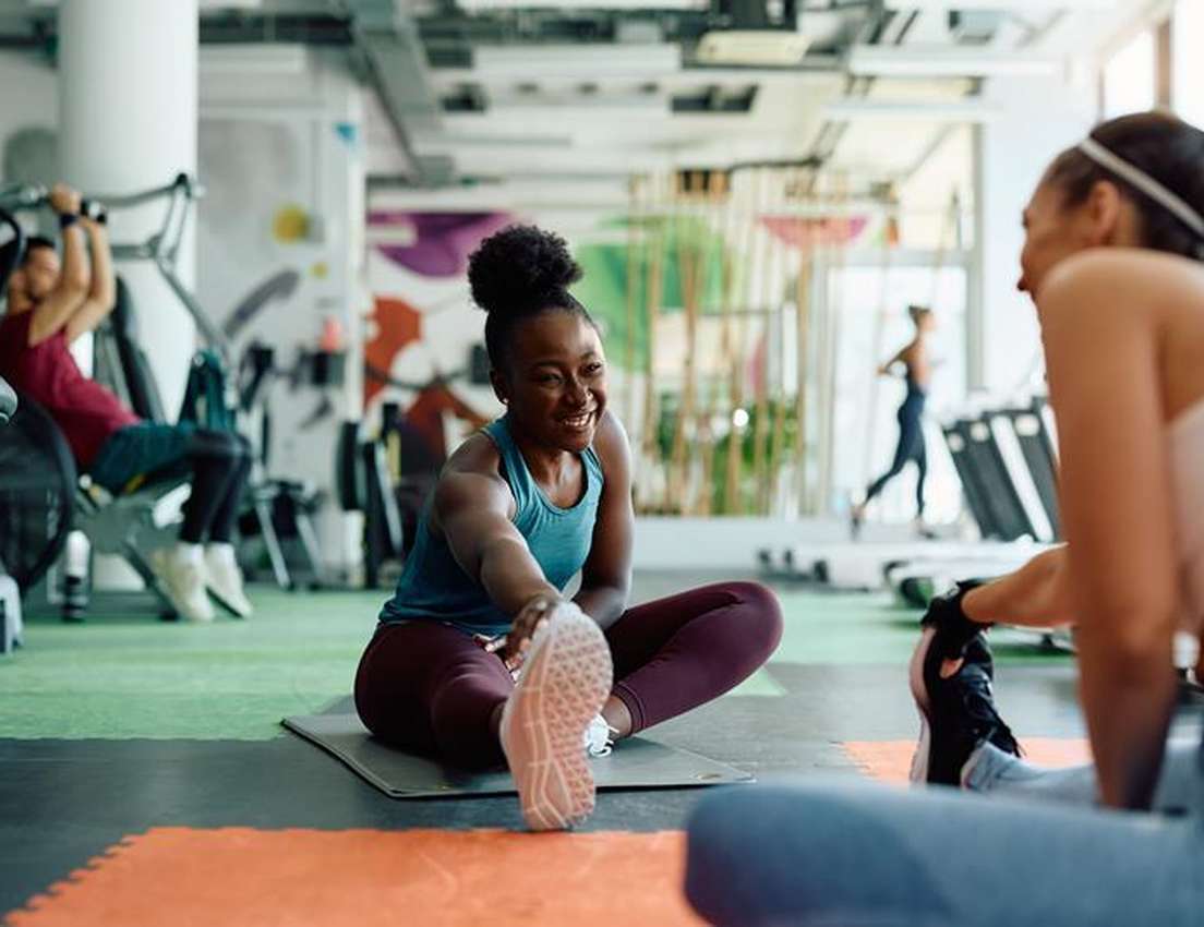 A smiling Black woman sits on a mat, stretching her leg forward in a vibrant gym with other exercisers around her.