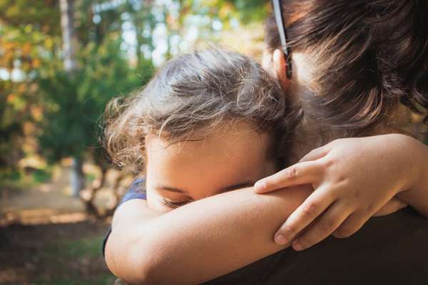 A child with eyes closed embraces an adult tightly, resting their head on the adult's shoulder outdoors.