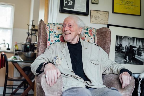 Elderly man with white hair smiling and looking left, sitting in a patterned armchair indoors. He wears a beige jacket over a