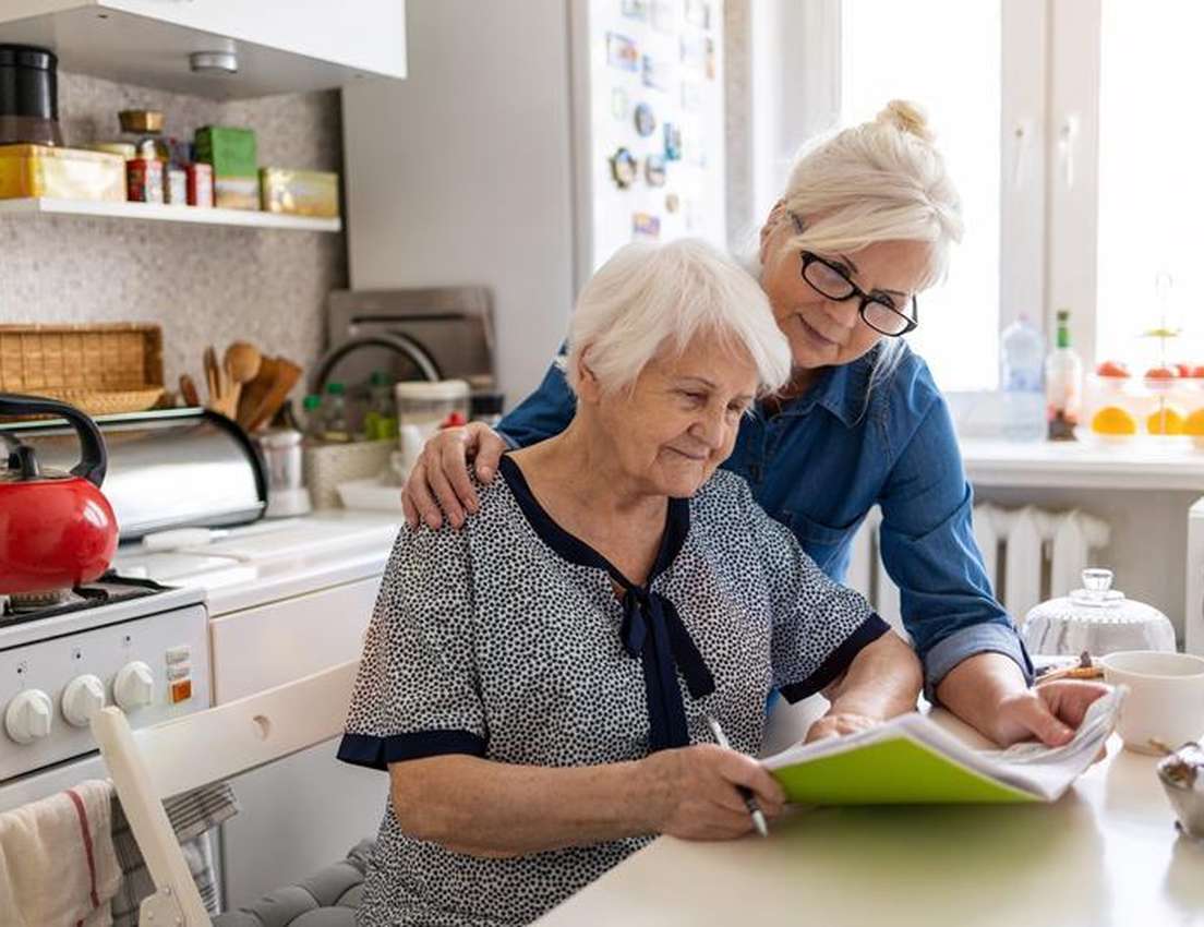 Two older women in a bright kitchen. One sits writing in a notebook, while the other stands behind her, looking on with a sup