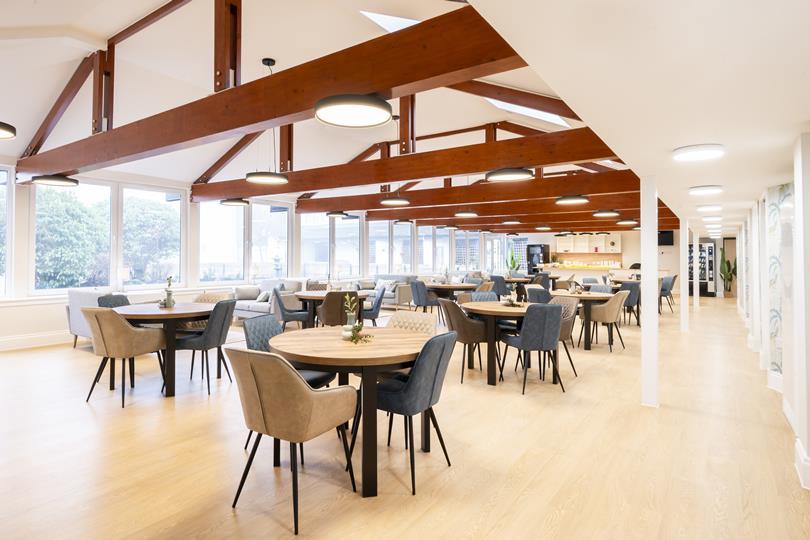 Spacious, sunlit dining hall with numerous round tables, upholstered chairs, exposed timber beams, and large windows overlooking trees.