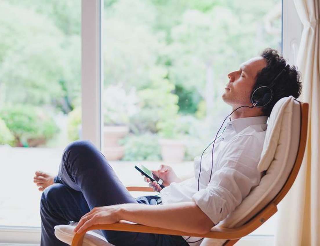 Man in a chair near a window, wearing headphones, eyes closed, holding a smartphone, and relaxing in a serene setting.