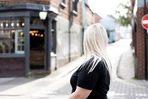 Back of a blonde woman in a black shirt, looking down a narrow, curving cobblestone street with a cafe on the left.