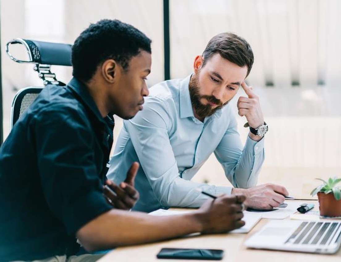Two male colleagues in an office. A Black man speaks and writes, while a white bearded man listens intently, hand on his temp