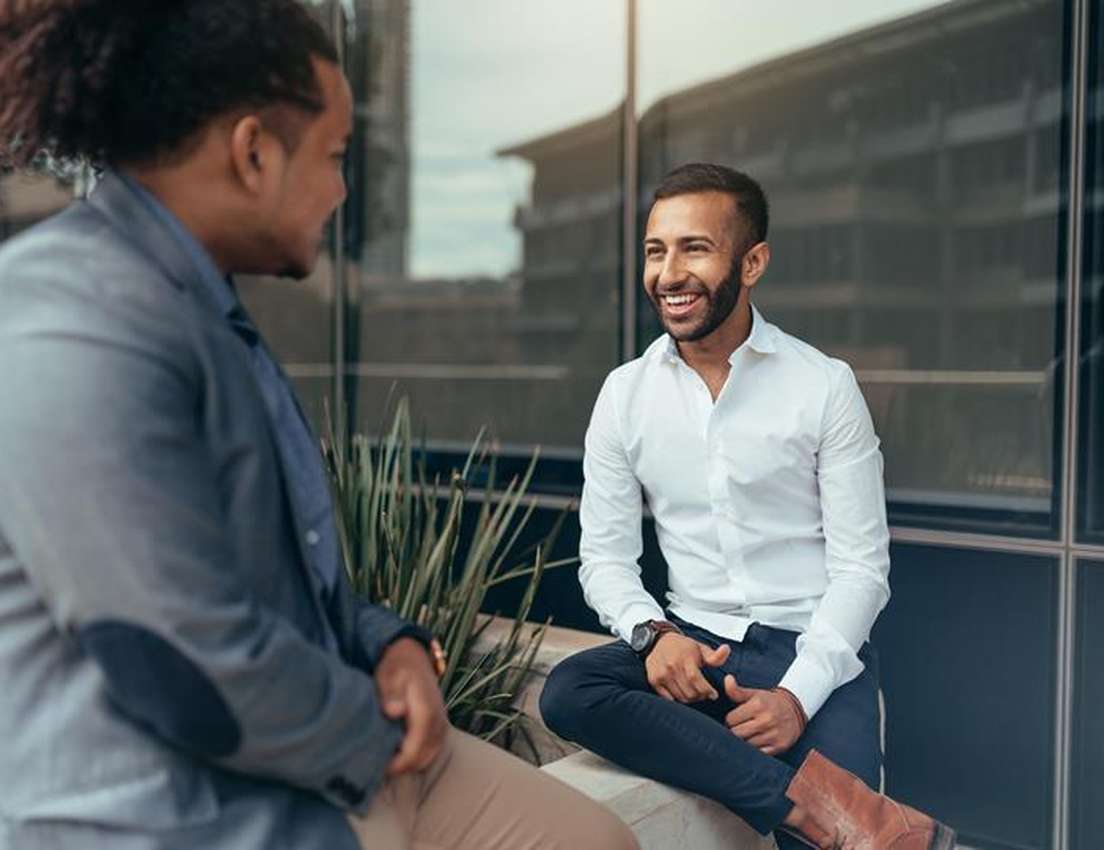 Two men in business attire are sitting outside a building, engaged in a lively conversation and smiling at each other.