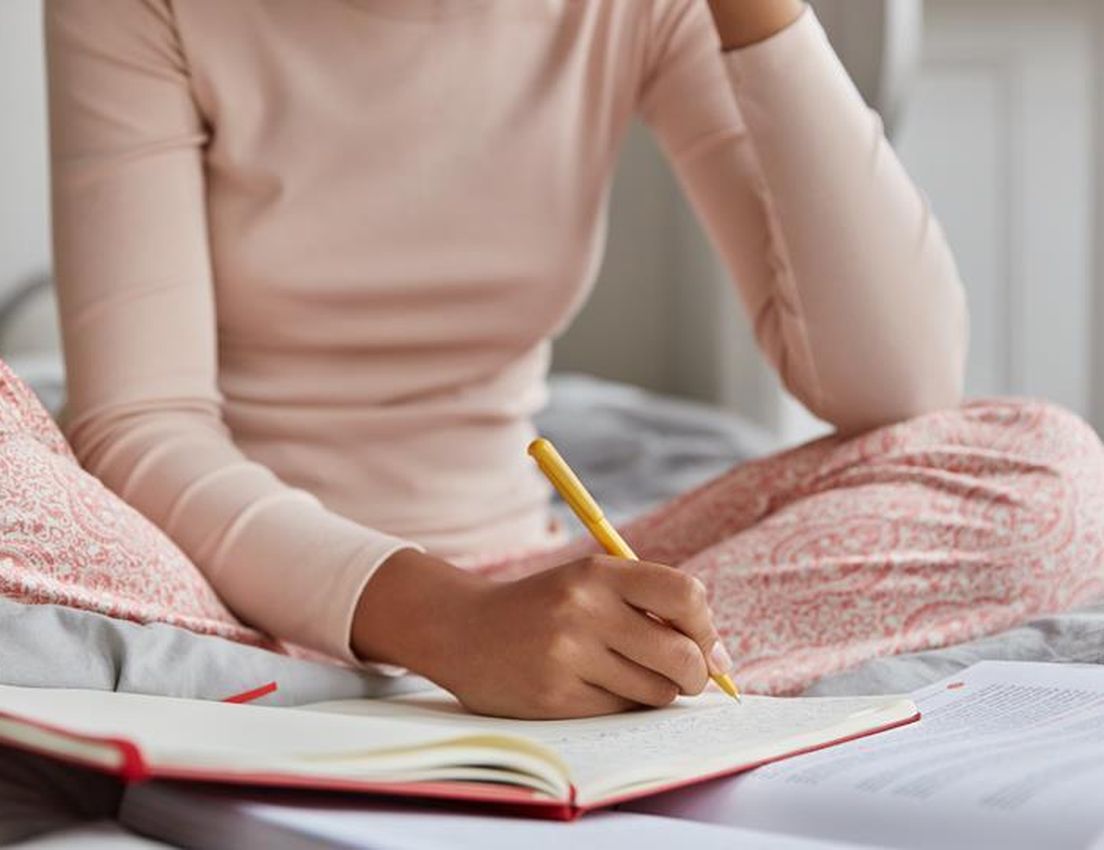 Person in pink pajamas sitting on a bed, writing in a notebook with a yellow pencil, surrounded by open books.