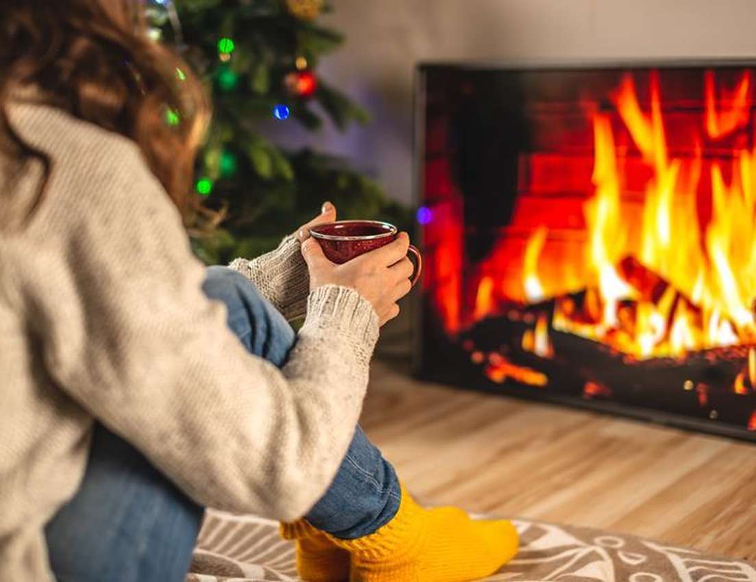 A person, from behind, holding a red mug, sits on a rug near a twinkling Christmas tree and a digital fireplace.