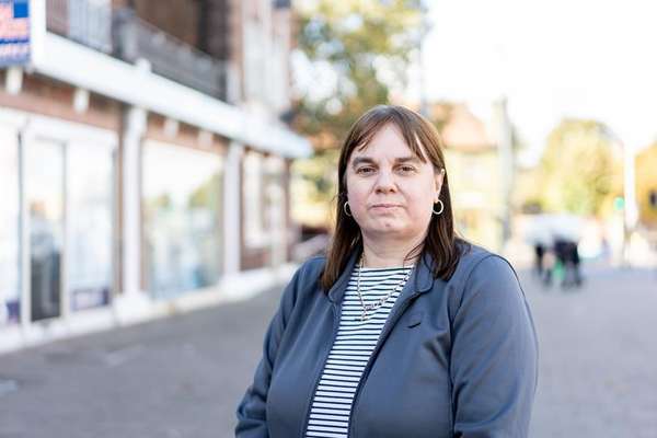A middle-aged woman with brown hair, bangs, gold hoop earrings, a striped shirt, and a grey jacket stands on a blurry urban street.