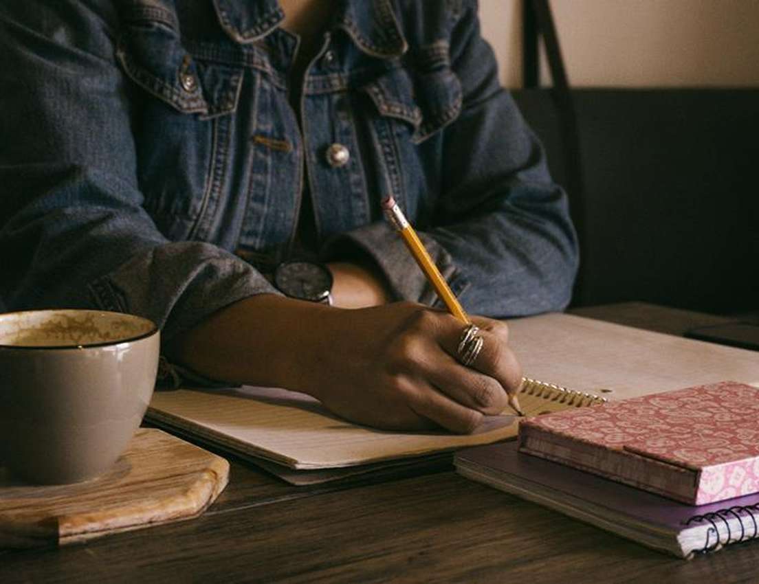A person in a denim jacket writes in a spiral notebook with a pencil, a coffee cup with foam on a wooden coaster beside them.