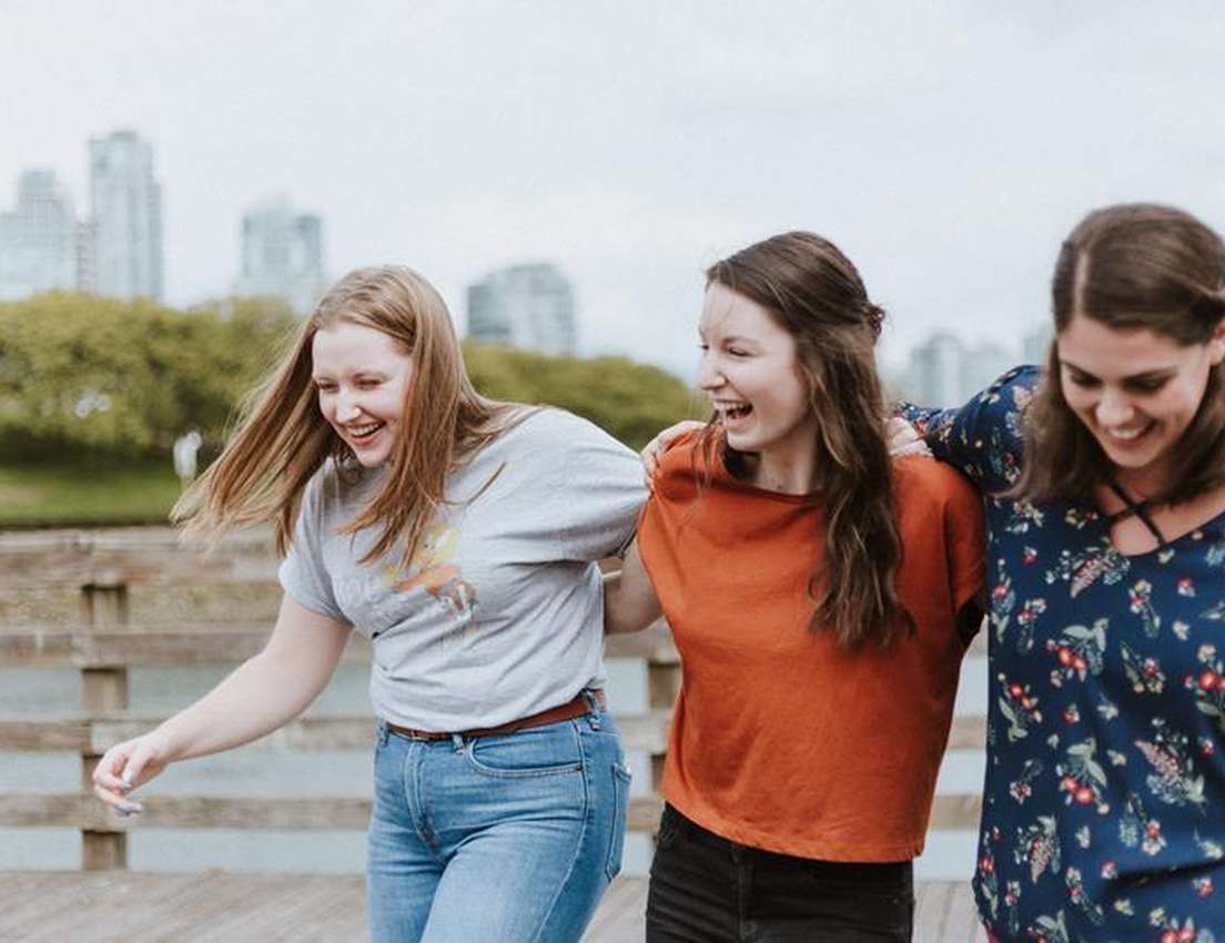 Three women smiling and laughing, walking arm-in-arm on a boardwalk with a city skyline in the background.