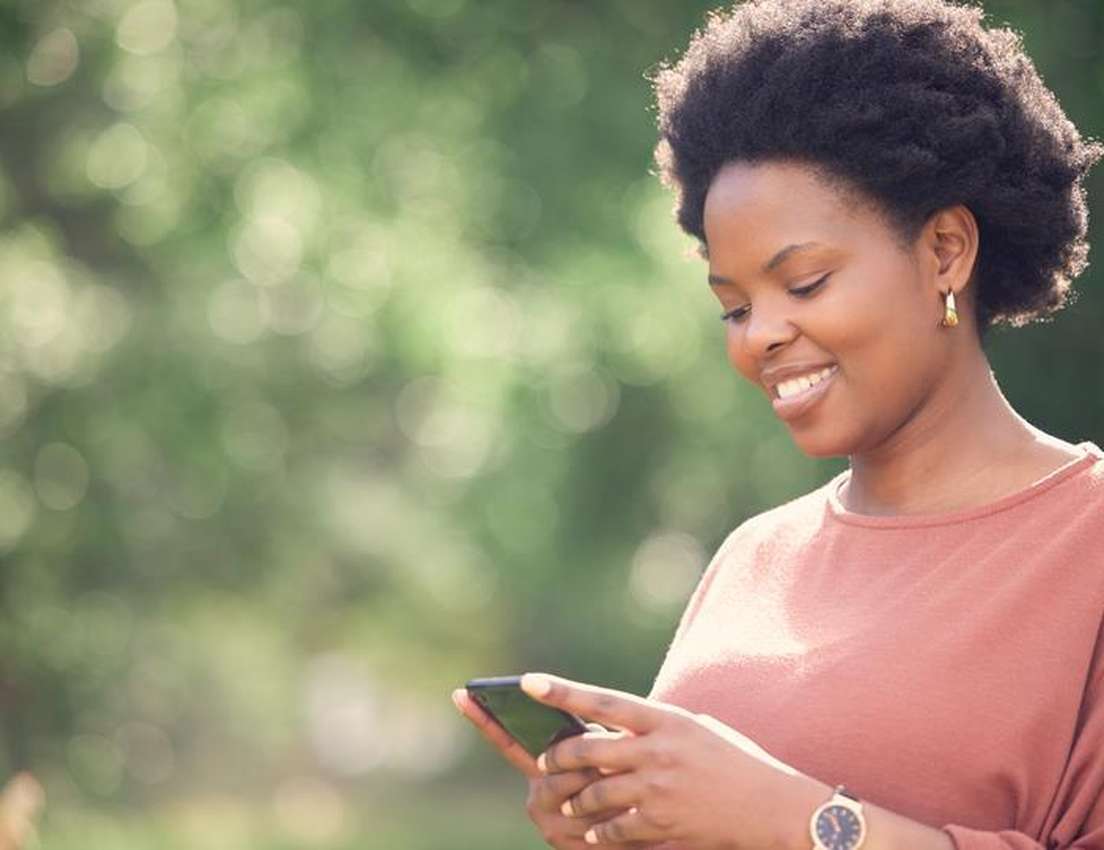 Smiling woman using a smartphone outdoors against a blurred green background.