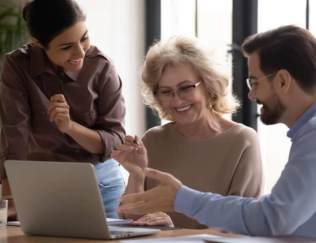 Three smiling, diverse colleagues, including an older woman, collaborate around a laptop.