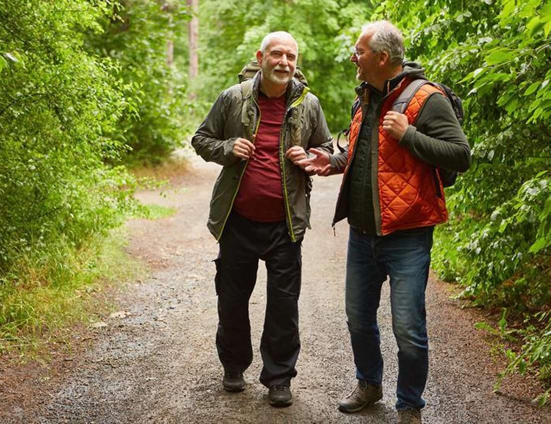 Two older men hiking on a forest path, wearing jackets and backpacks, smiling and engaged in conversation surrounded by lush 