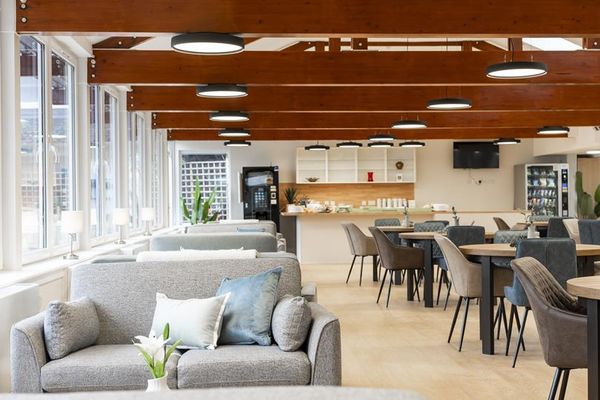 Bright modern lounge with grey sofas, dining tables, and a counter area. Wooden beams on the ceiling with circular lights.