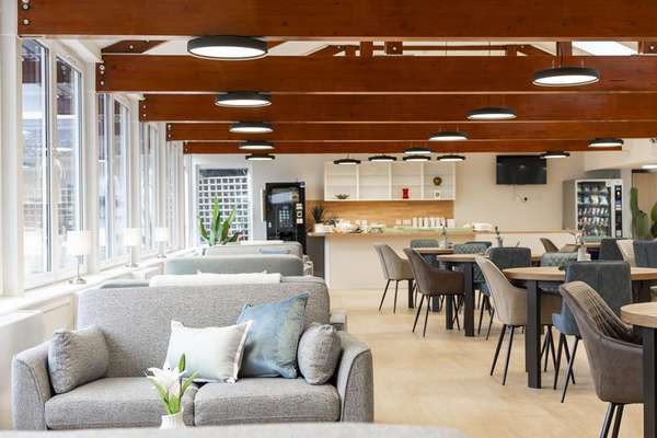 Bright modern lounge with grey sofas, dining tables, and a counter area. Wooden beams on the ceiling with circular lights.