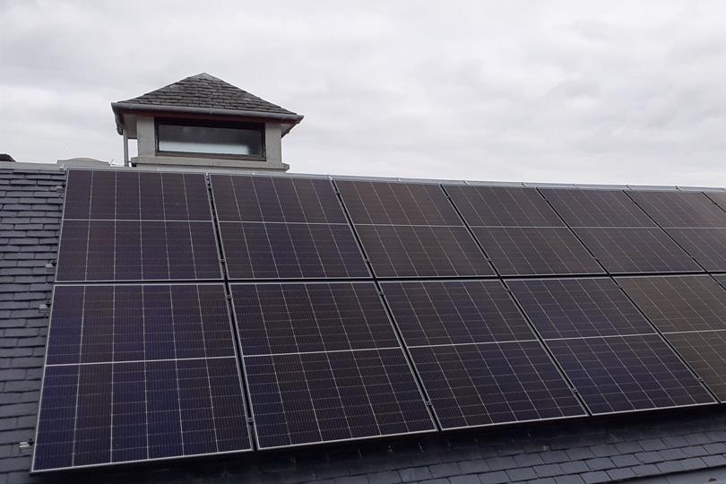 Numerous black solar panels cover a dark gray slate roof, with a small cupola and a cloudy sky in the background.