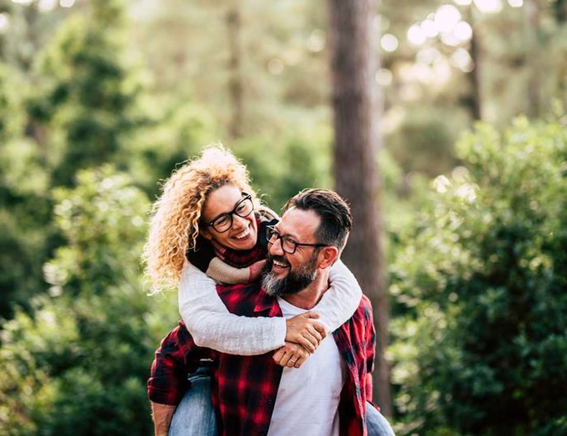 Happy woman with curly hair riding piggyback on a bearded man in a sunny forest, both wearing glasses and smiling.