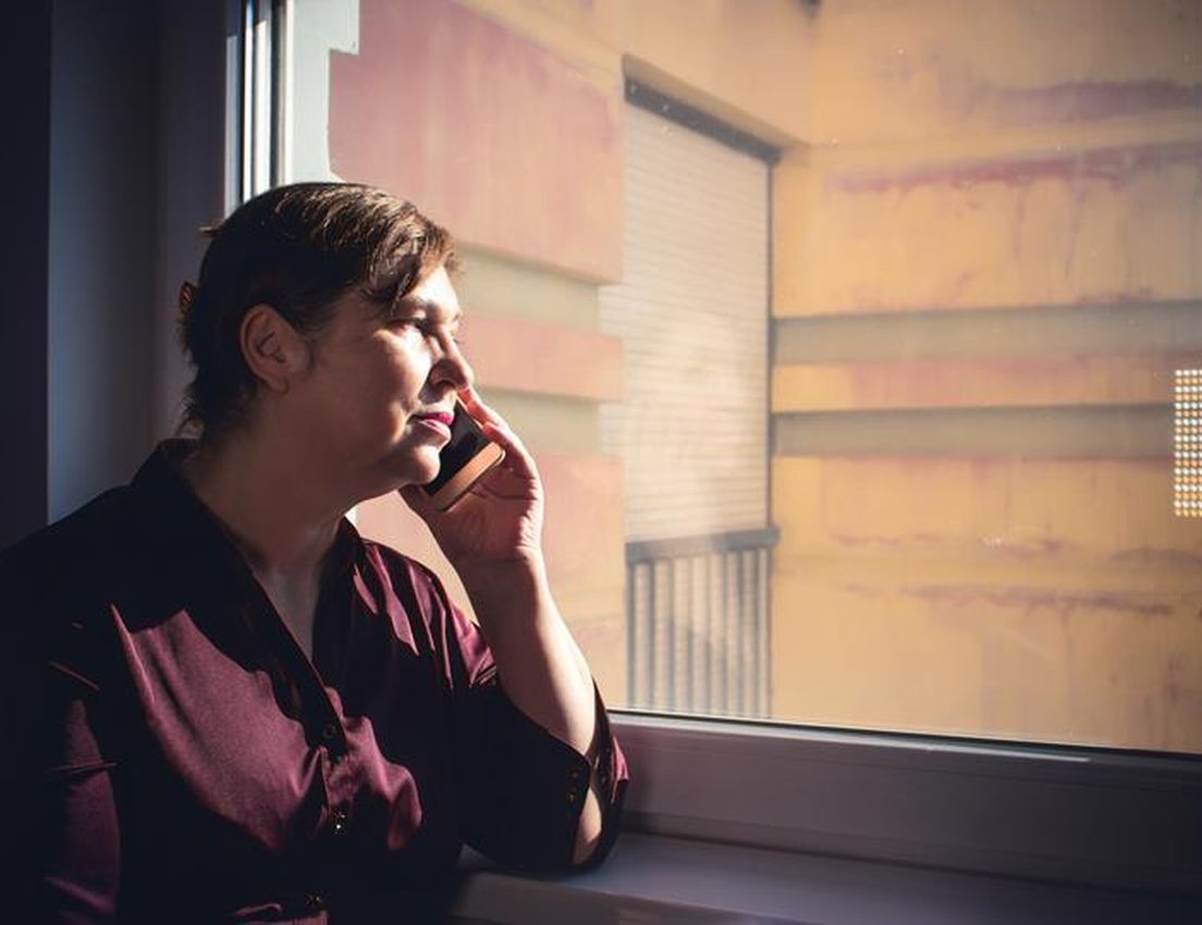 Woman talking on a cell phone by a window, sunlight illuminating her face as she looks out at a building.