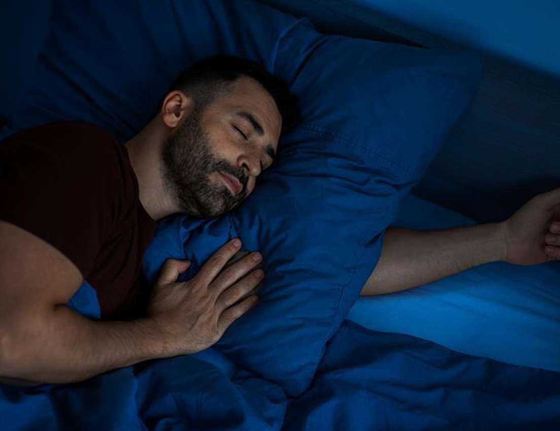 A man sleeps peacefully in bed, resting his head on a blue pillow. He's covered with a blue blanket, wearing a dark shirt.
