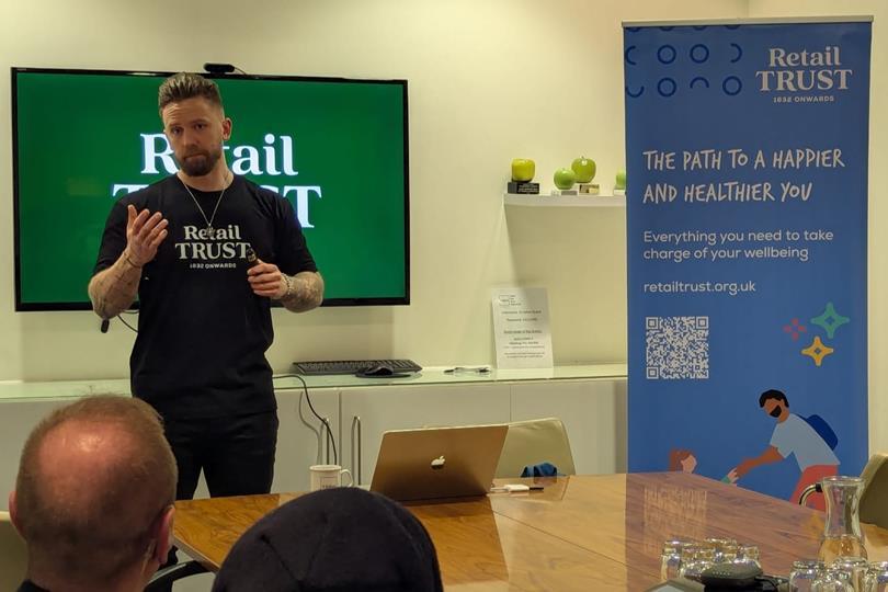 A man in a "Retail Trust" t-shirt speaks at a presentation, with "Retail Trust" branding on a screen and a wellbeing banner.