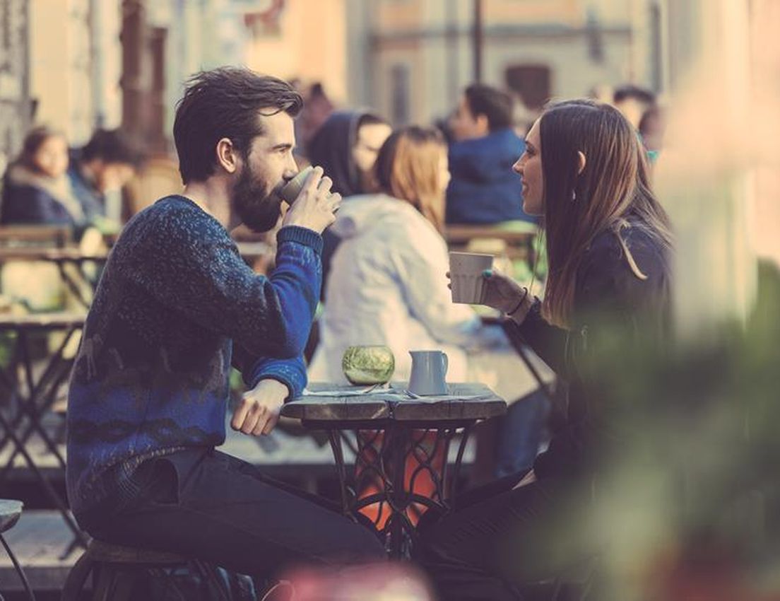 Two people, a man with a beard and a woman, enjoy coffee and conversation at a rustic outdoor cafe table on a sunny day.