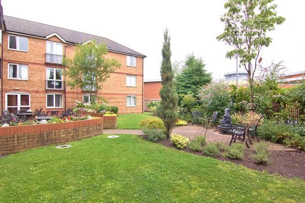 A brick apartment building overlooks a landscaped garden with a manicured lawn, flower beds, a small tree, and a seating area