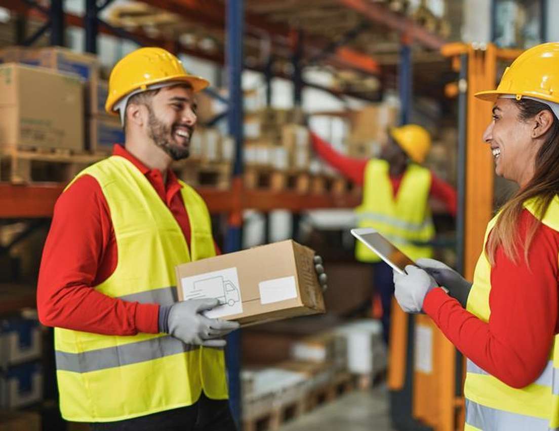 Two smiling warehouse workers in hard hats and vests, one holding a package and the other a tablet, collaborating in a wareho