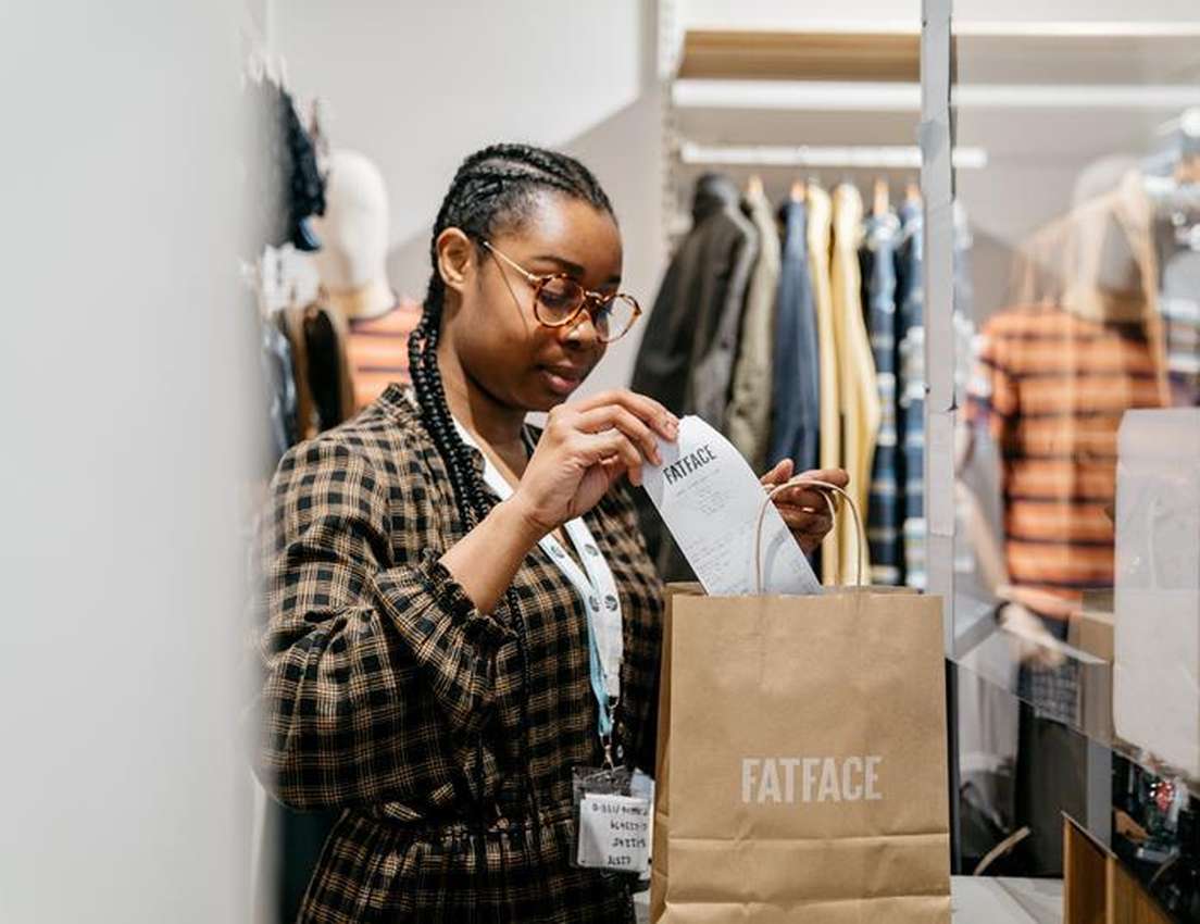 A young woman with braided hair and glasses, wearing a plaid shirt, places a receipt into a brown "FATFACE" shopping bag in a