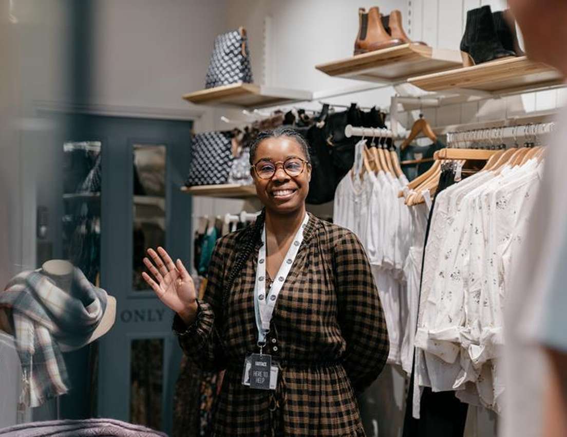 Smiling Black woman in glasses and plaid dress, wearing "HERE TO HELP" tag, waves in a clothing store.