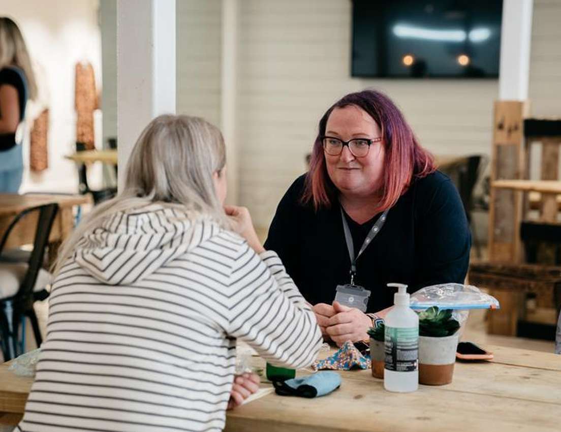 Two people seated at a wooden table in conversation. One wears a striped hoodie, the other has pink hair. Hand sanitizer is o