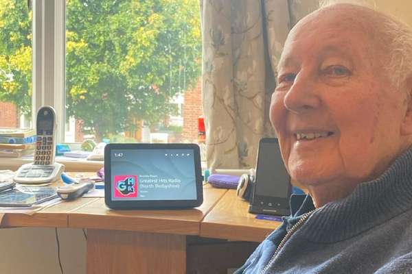 An elderly man smiles at the camera, sitting at a wooden table with a smart display showing "Greatest Hits Radio" by a window.