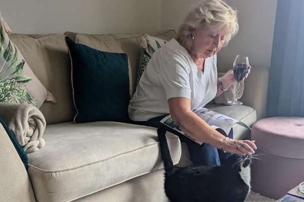 Blonde woman on a couch sips red wine, reads a magazine, and pets a black and white cat looking up at her.