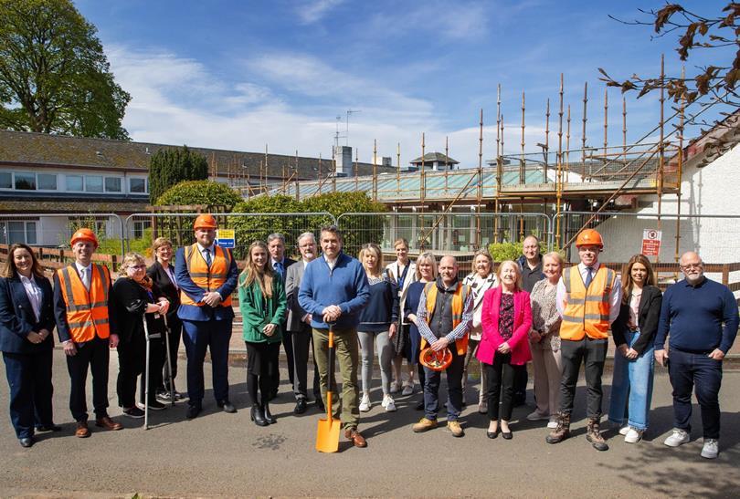 A group of people, some in hard hats and vests, pose with a shovel in front of a building with scaffolding, signifying a groundbreaking.