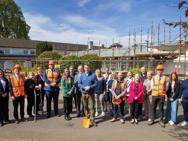 A group of people, some in hard hats and vests, pose with a shovel in front of a building with scaffolding, signifying a groundbreaking.