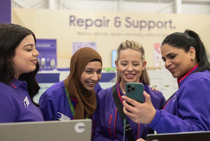 Four diverse women in purple tops smile while looking at a smartphone, with a "Repair & Support" sign in the background.