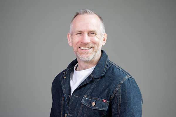 Smiling man with gray hair and a short beard, wearing a dark blue denim jacket over a white t-shirt, smiling at the camera.