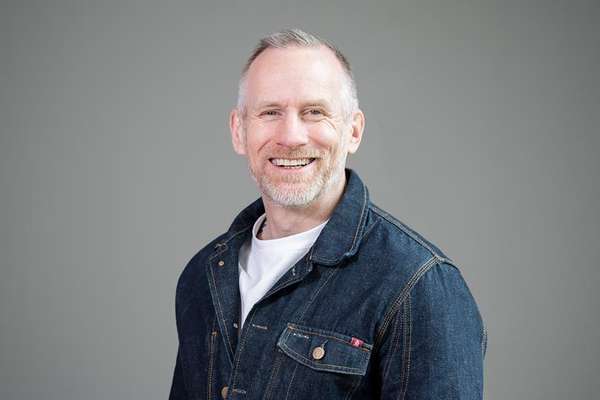 Smiling man with gray hair and a short beard, wearing a dark blue denim jacket over a white t-shirt, smiling at the camera.