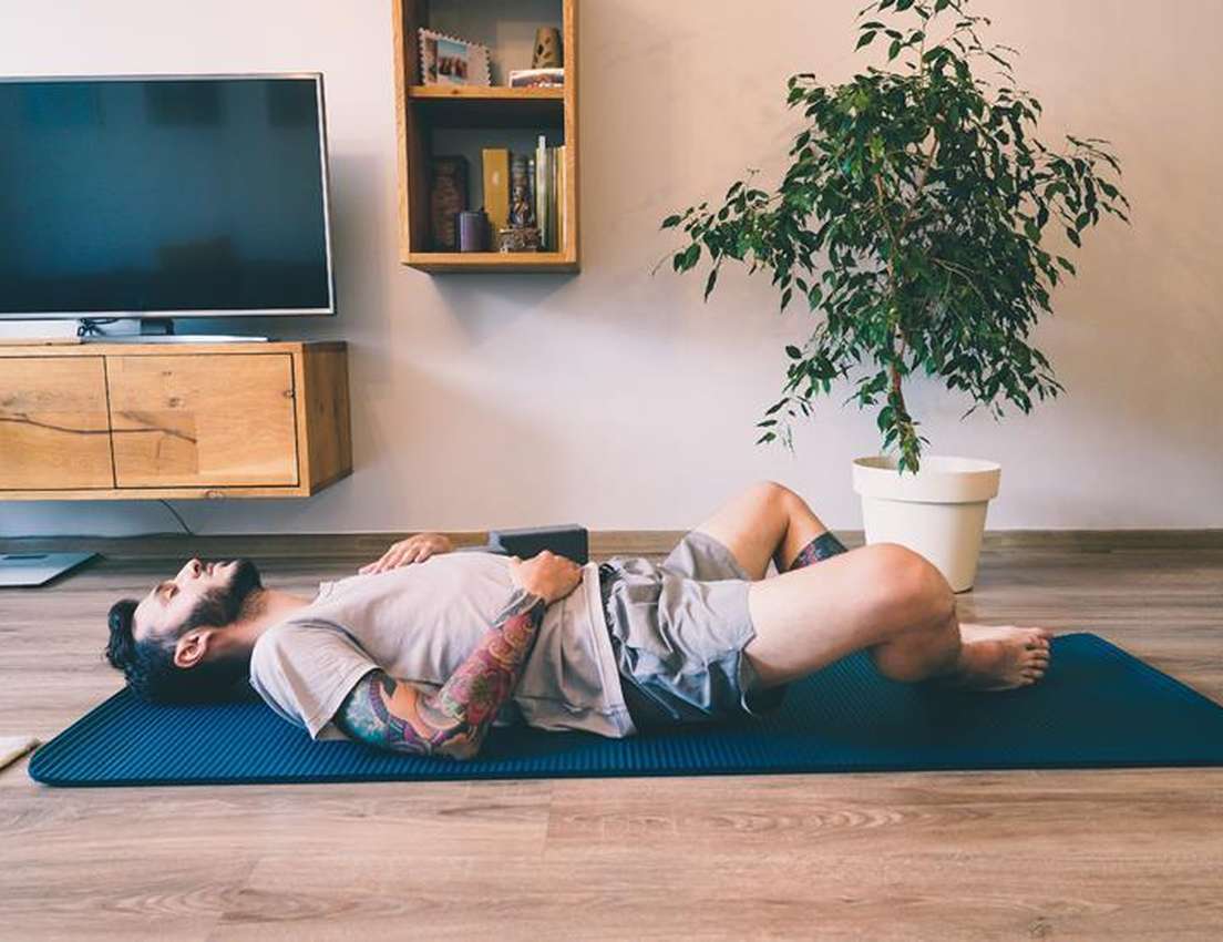 Man lying on a yoga mat at home, with legs bent and hands on stomach, in a room with a TV, wooden cabinet, and potted plant.
