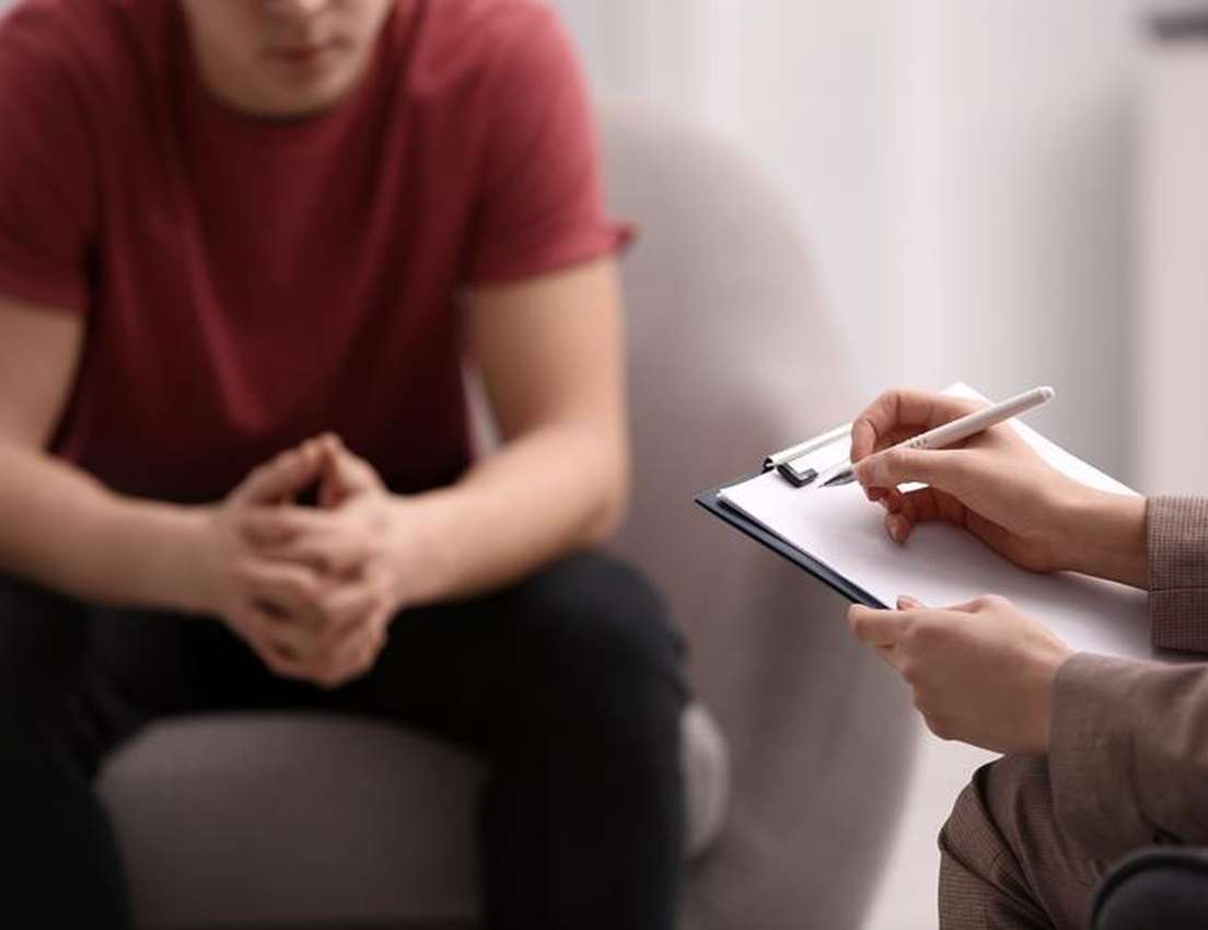 Person in a red shirt sits opposite another with a clipboard and pen, possibly in a counseling or therapy session.