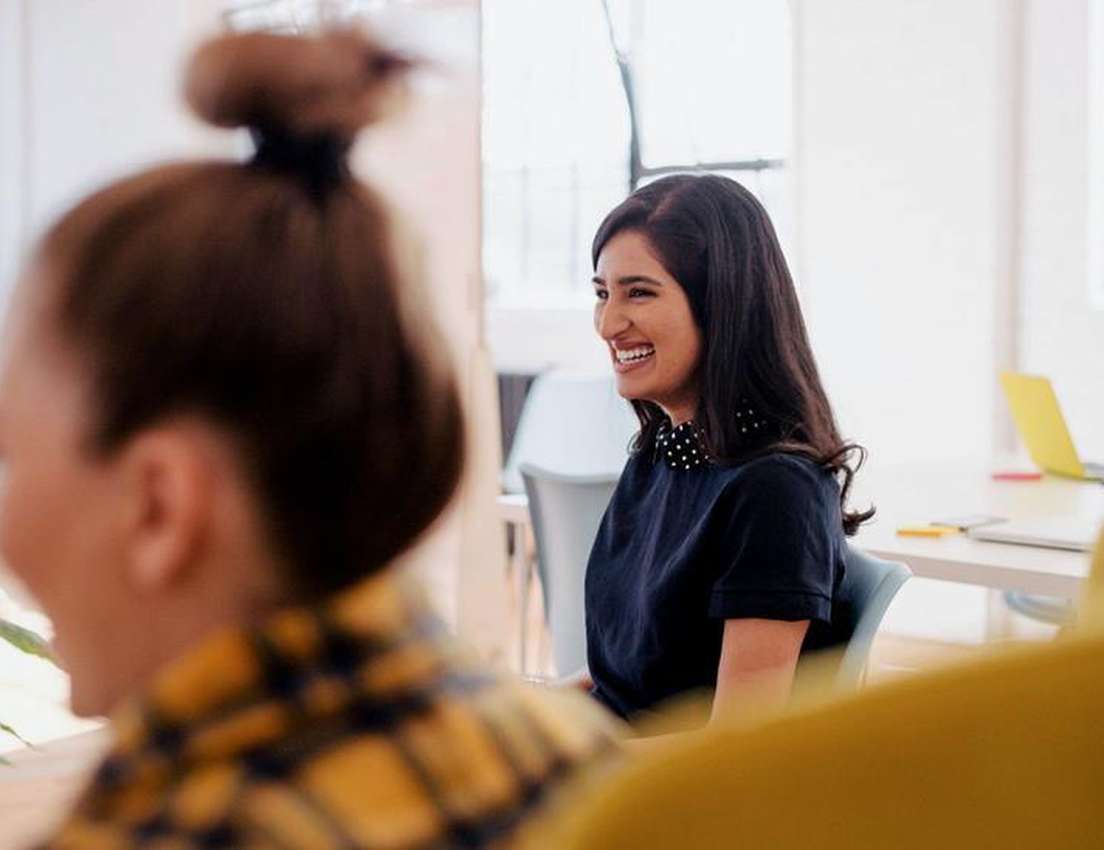 A joyful woman with long dark hair and a polka-dot collar smiles, sitting in an office meeting. A blurred person is in the fo