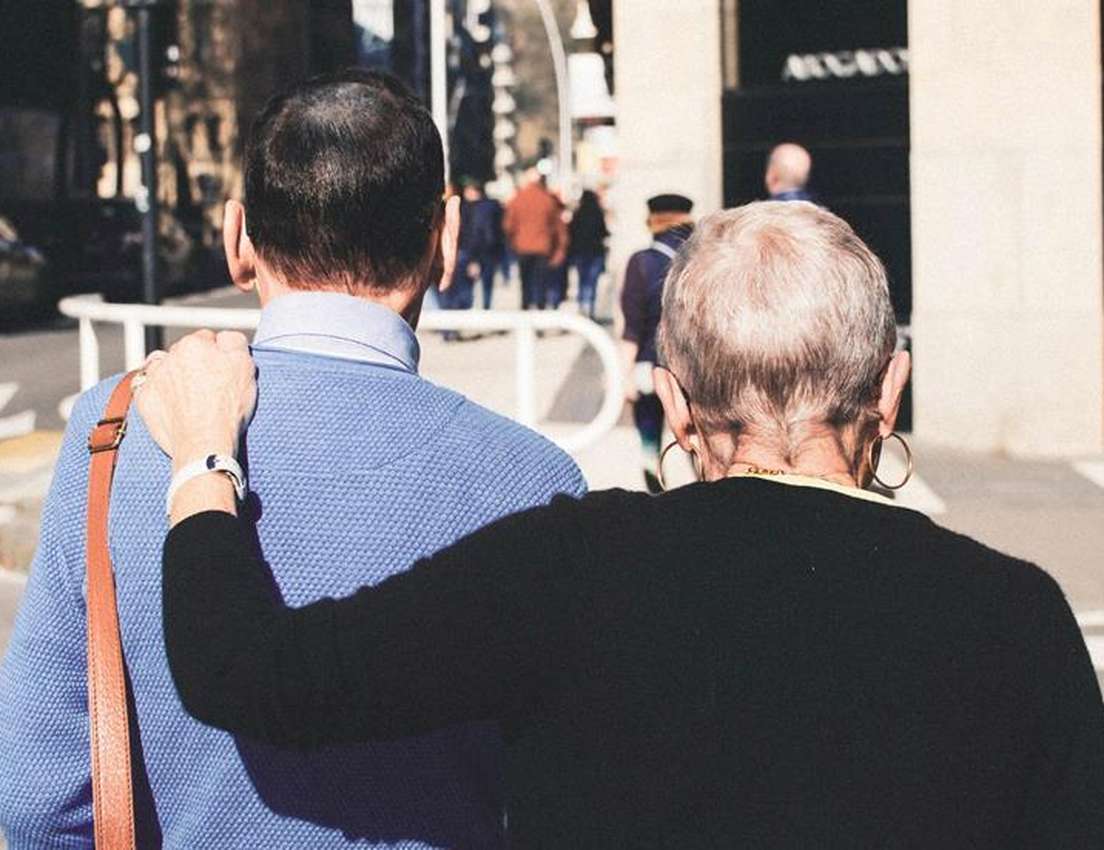 Rear view of two people walking: a grey-haired person with hoop earrings has an arm around a dark-haired person in a blue swe