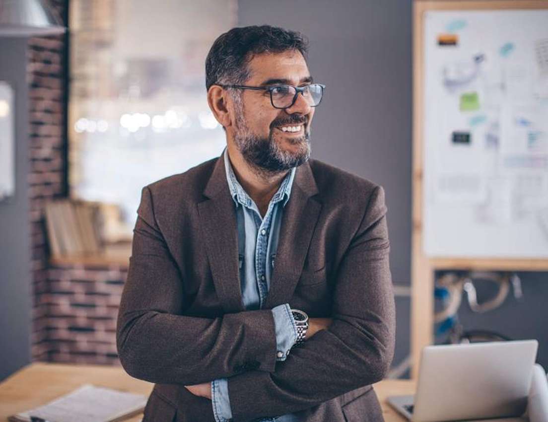 Man with glasses and a beard, wearing a suit jacket, smiles while standing with crossed arms in an office setting with a lapt
