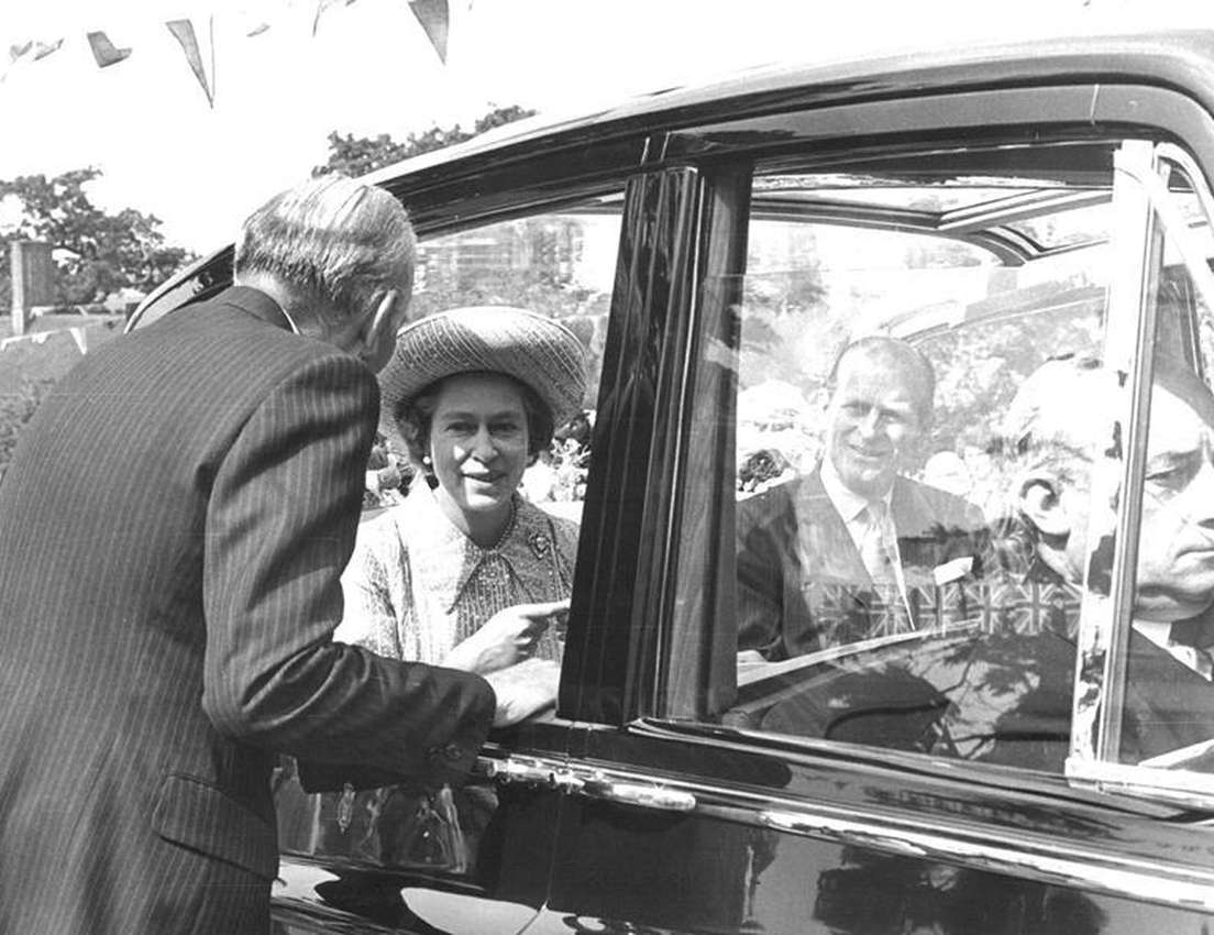 Black and white photo. Queen Elizabeth II, in a car, smiles and points while speaking to a man. Prince Philip smiles from ins