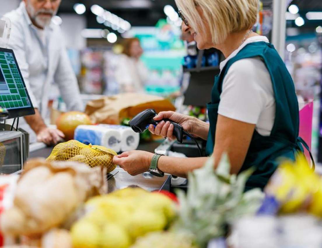Female cashier in green apron scans a bag of potatoes at a busy supermarket checkout. A male customer waits behind groceries.