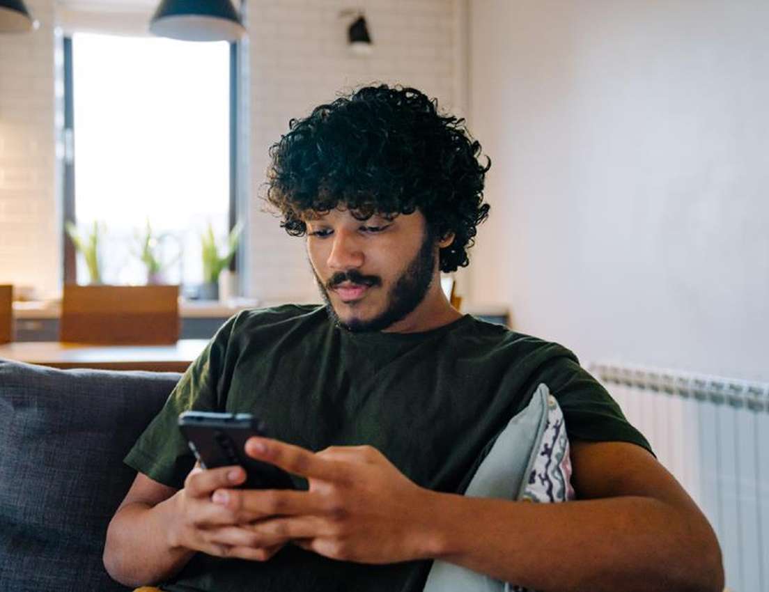 Young man with curly hair and a beard sits on a couch, intently looking at his smartphone.