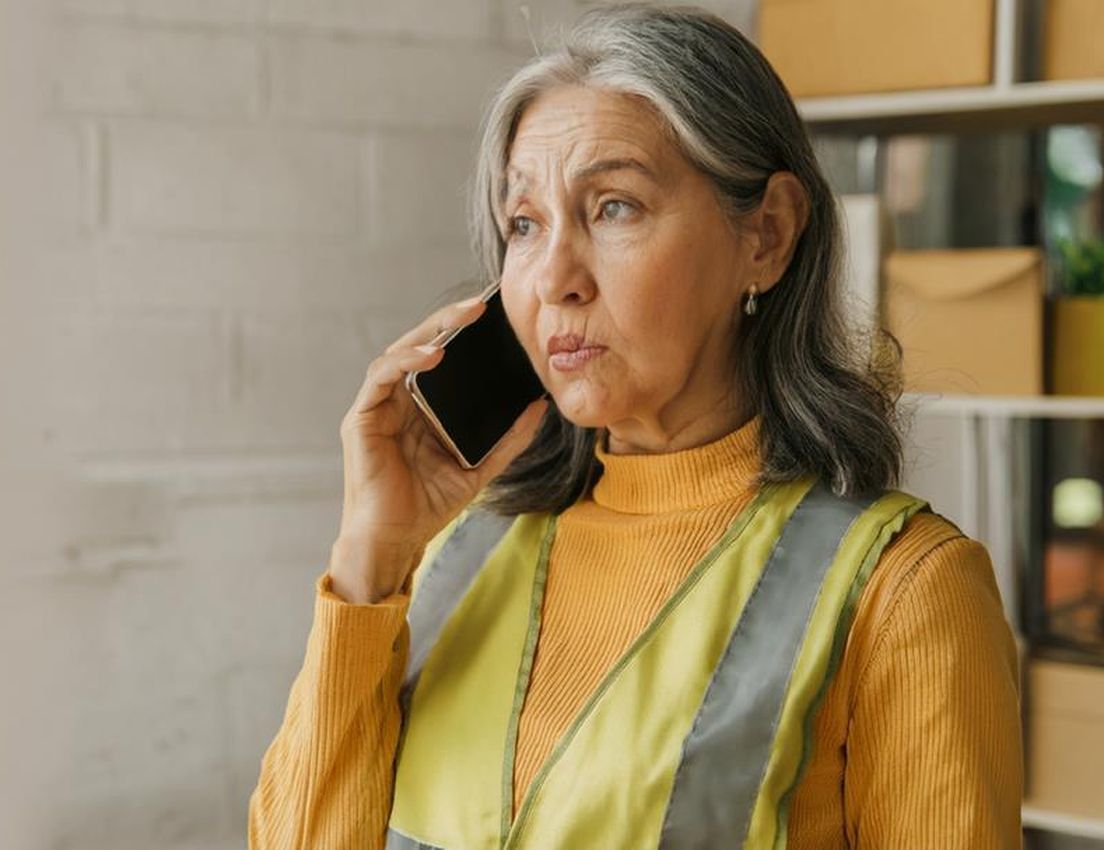 An older woman with long gray hair in a yellow turtleneck and reflective vest talks on a phone, looking pensive.
