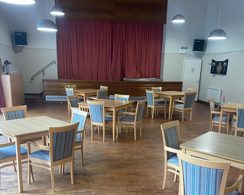 A community hall with a wooden floor, multiple light wooden tables, and chairs with blue striped cushions. A stage with red c