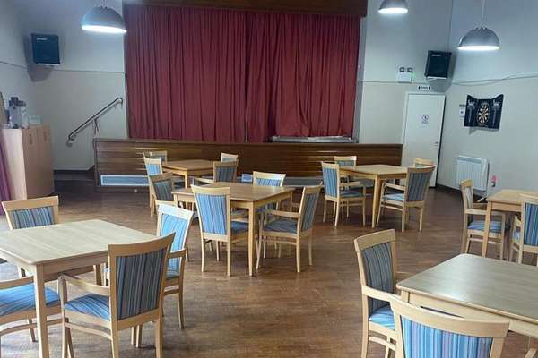 A community hall with a wooden floor, multiple light wooden tables, and chairs with blue striped cushions. A stage with red curtains is at the back.
