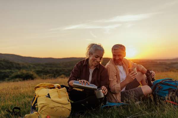 A smiling man and woman sit in a grassy field at sunset, sharing a warm drink from a thermos, with backpacks nearby.